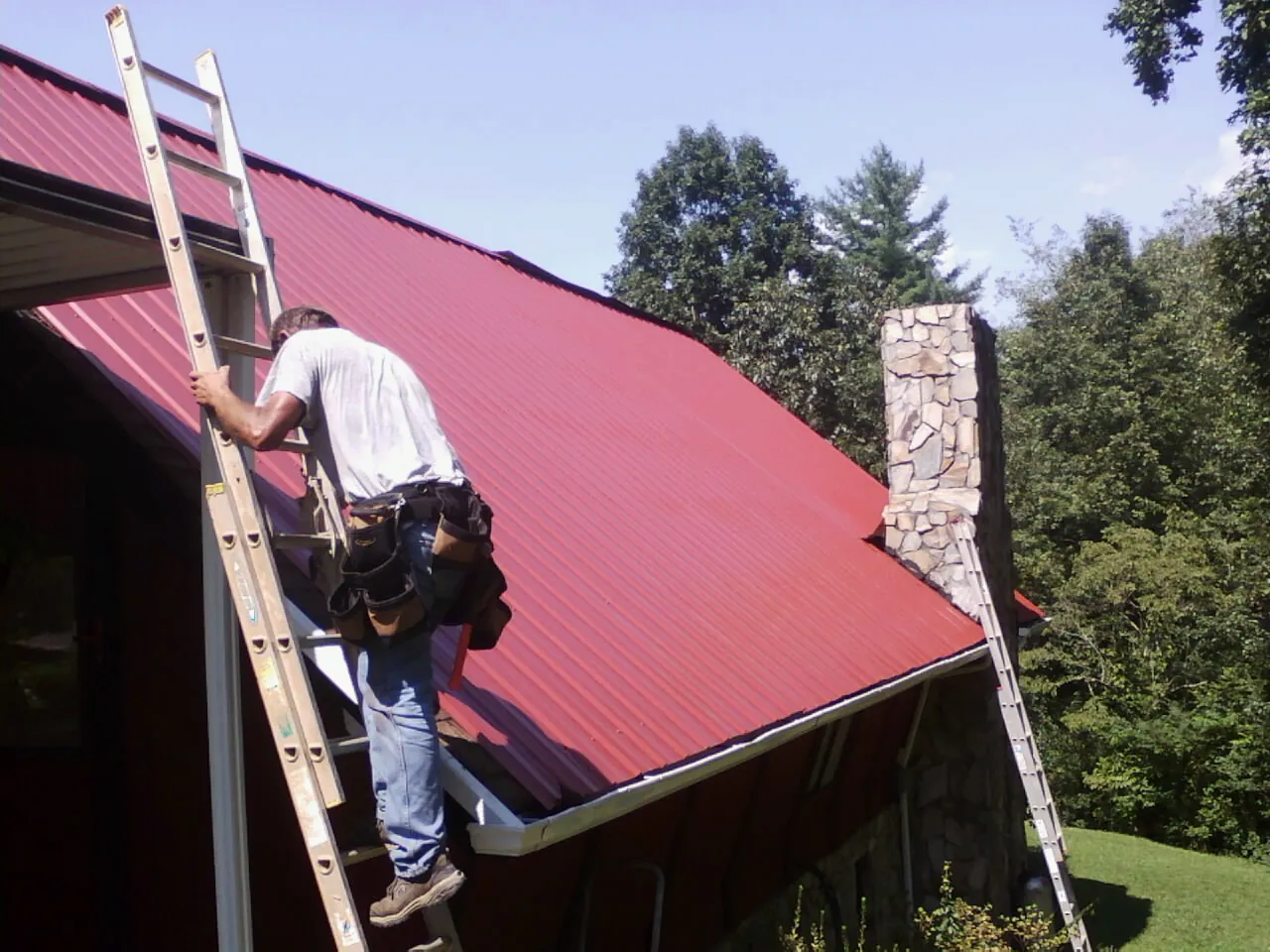 Roofer on a ladder beside a red metal roof and stone chimney — Soffit & Fascia Repair by Roofing Brothers in Falls Village